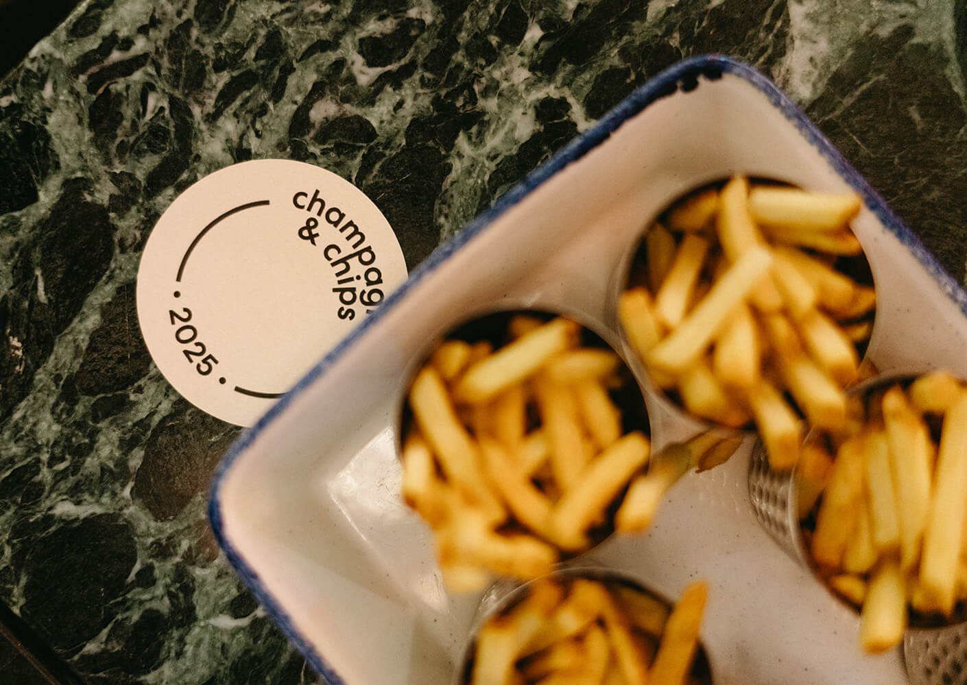 Fries in metallic cups with a 'champagne & chips 2025' coaster on a marble table