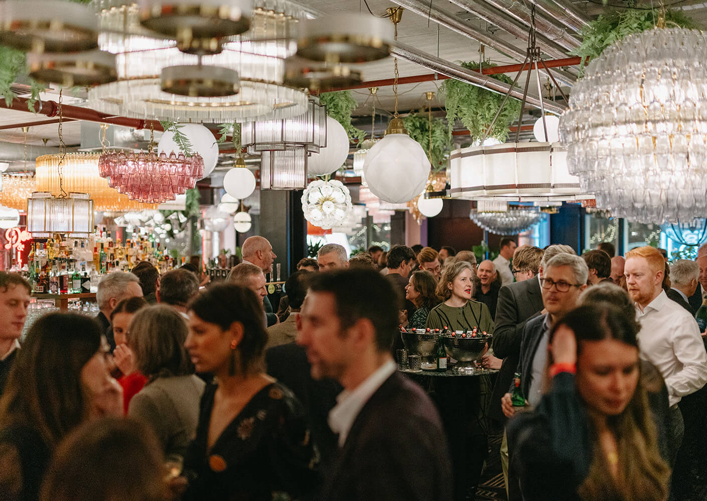 Crowded bar with people socializing under ornate chandeliers and warm lighting.