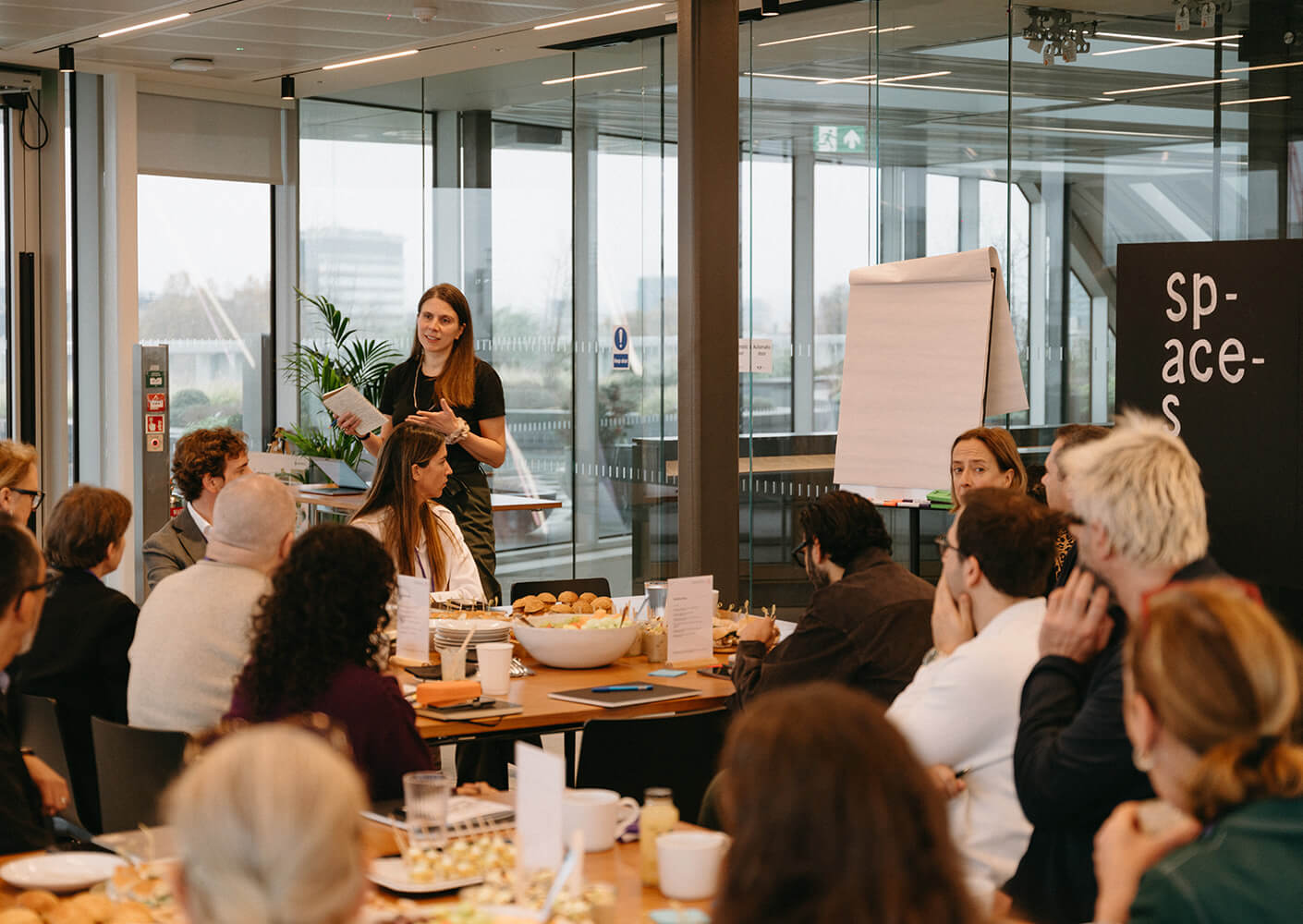 A group meeting in a well-lit room, with a speaker presenting to an engaged audience at a conference table filled with refreshments.