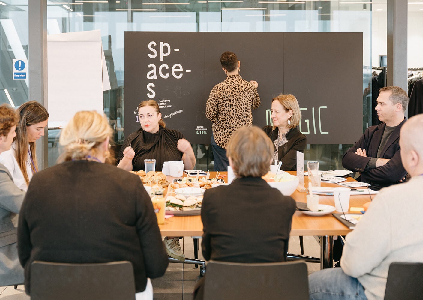 Group meeting at a table with food, discussing collaborative ideas. A speaker presents to attentive participants in a well-lit room.