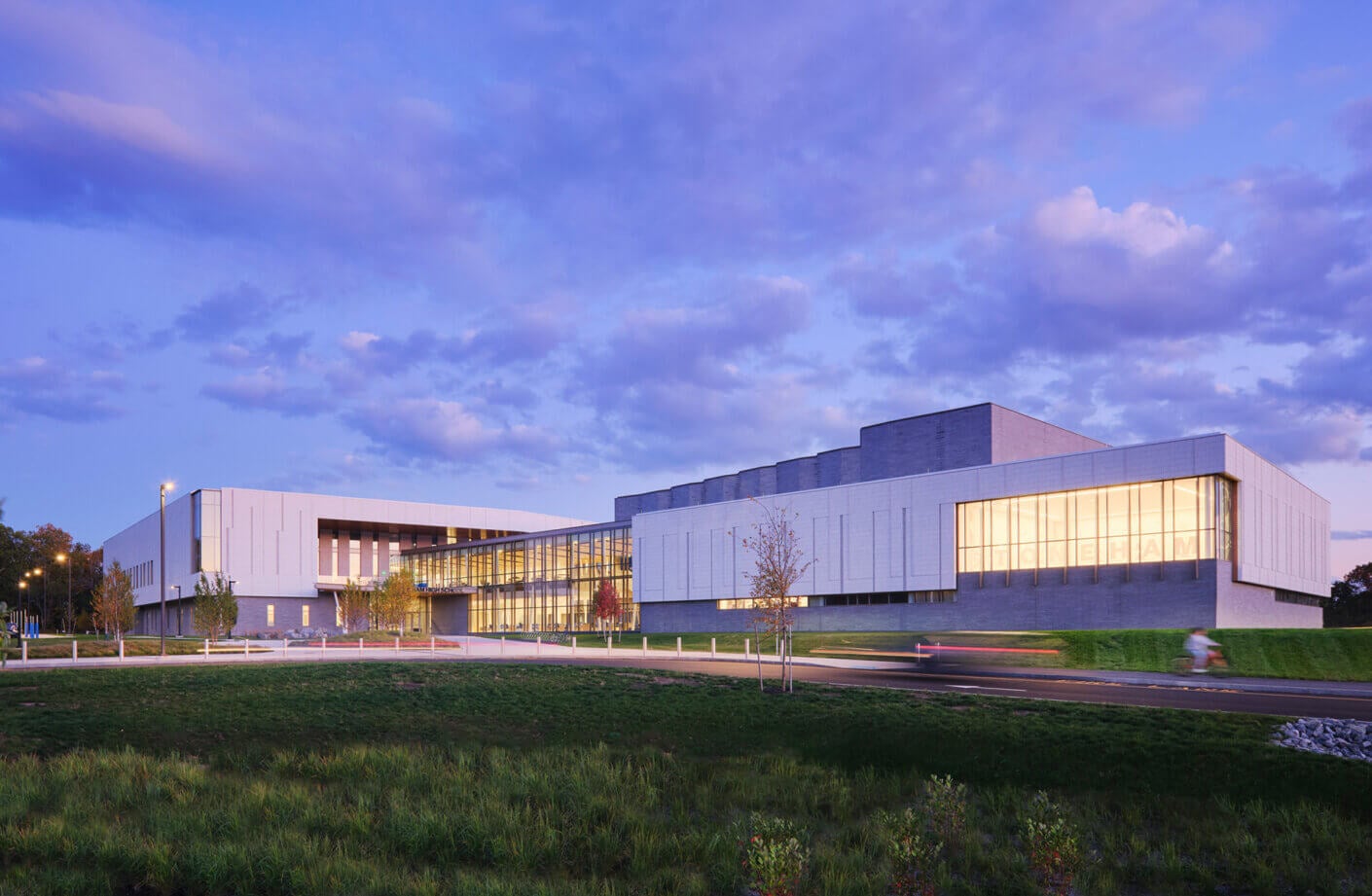 Architectural view of a school at dusk, showcasing large glass windows and landscaped surroundings, with a road in the foreground and cloudy sky.