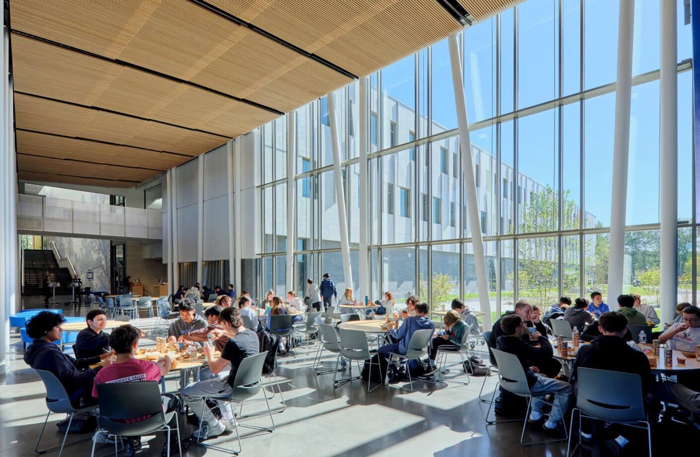 Students gather for lunch in a spacious cafeteria with large windows, wooden ceilings, and a bright, inviting atmosphere.