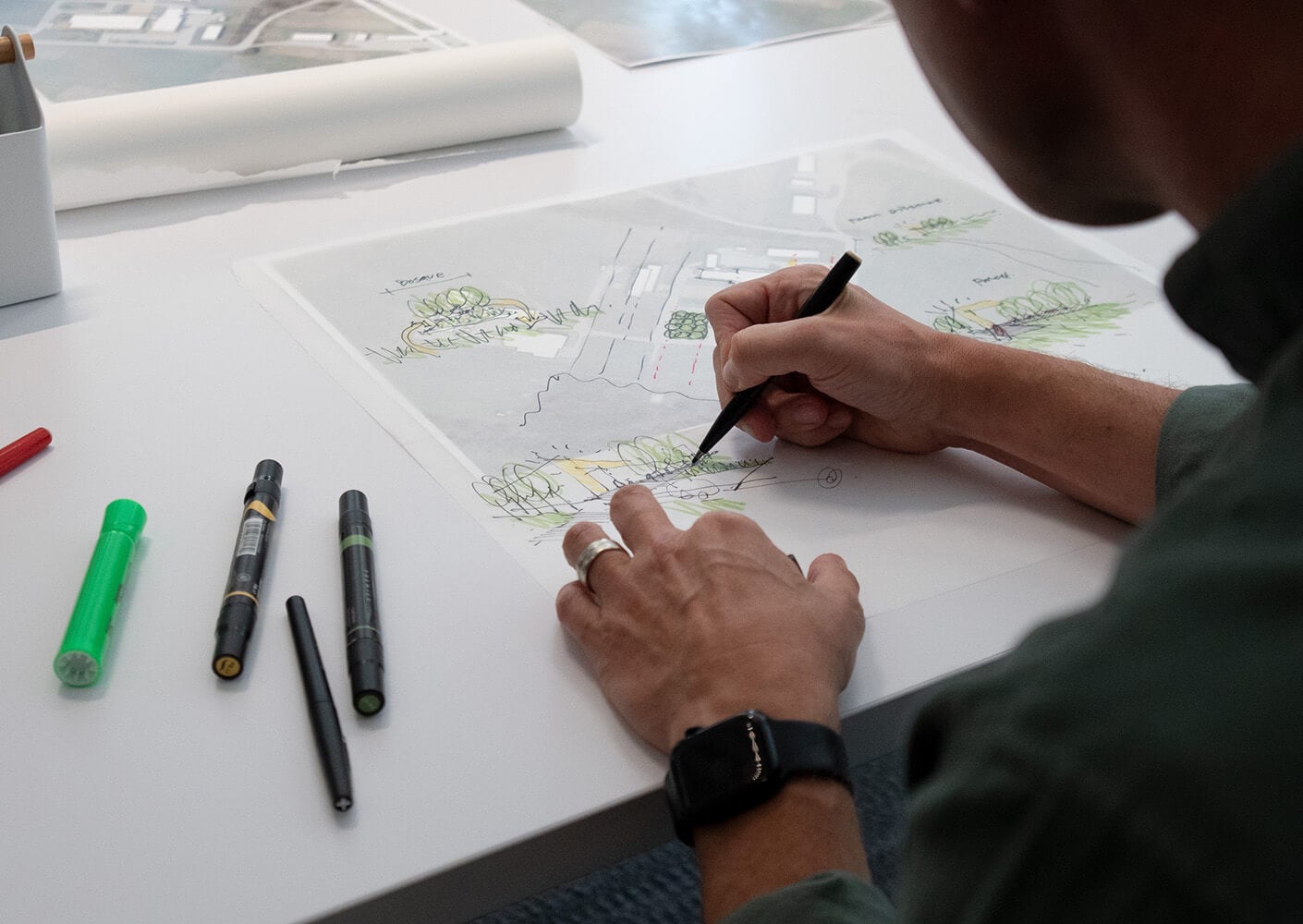 A person sketches a landscape design with colored markers on a clear sheet, surrounded by additional design materials on a table.