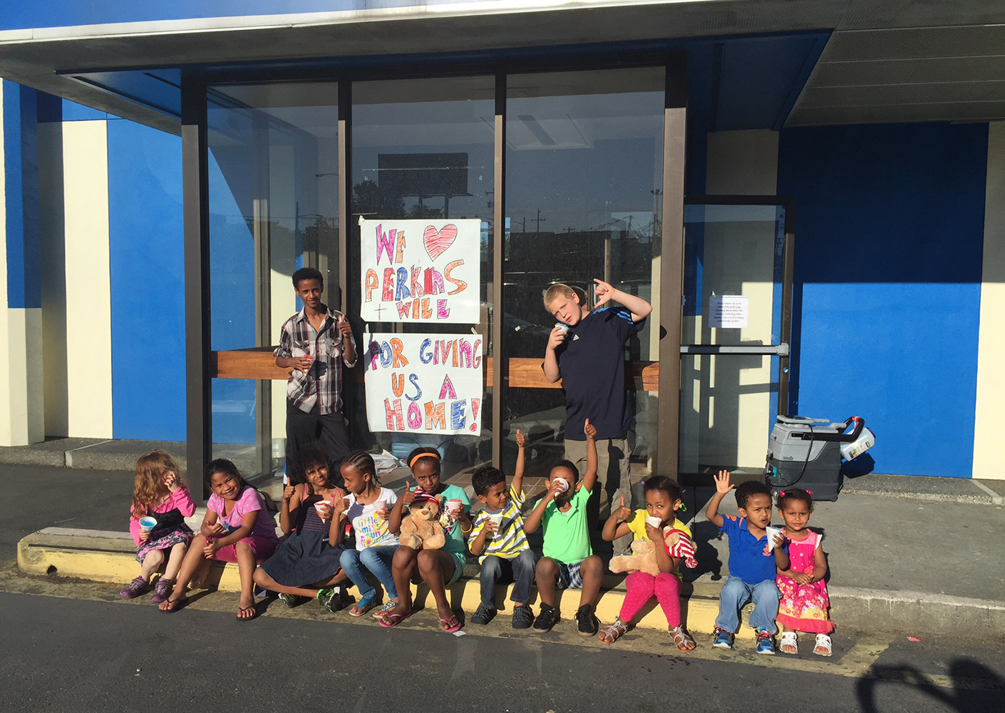 Kids gathered in front of building with hand-made sign that reads we love Perkins+Will
