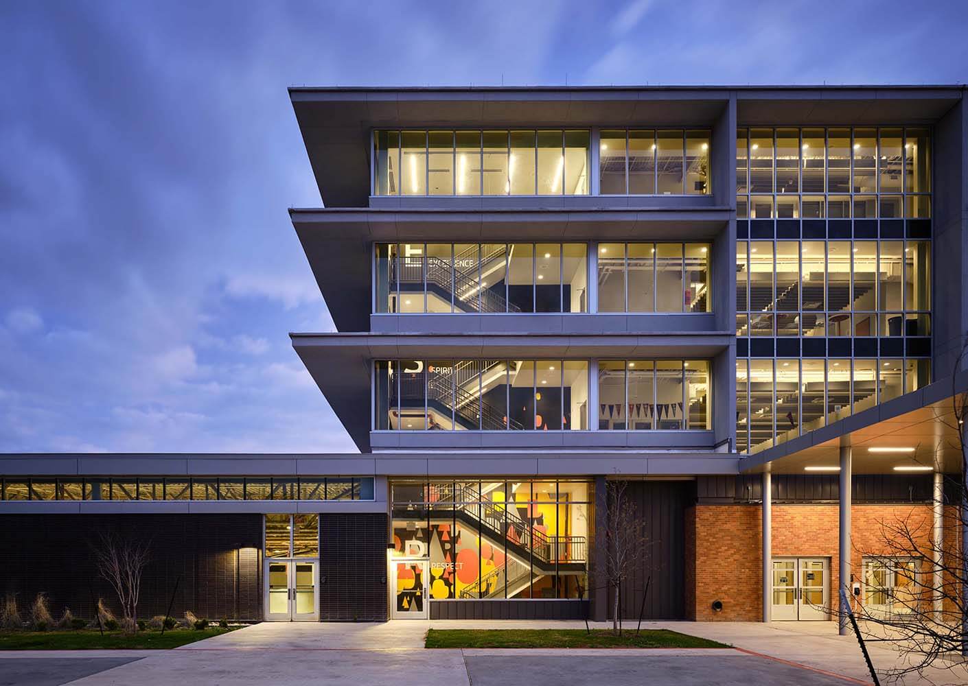 Modern multi-story school building exterior at dusk with illuminated windows and geometric design elements.