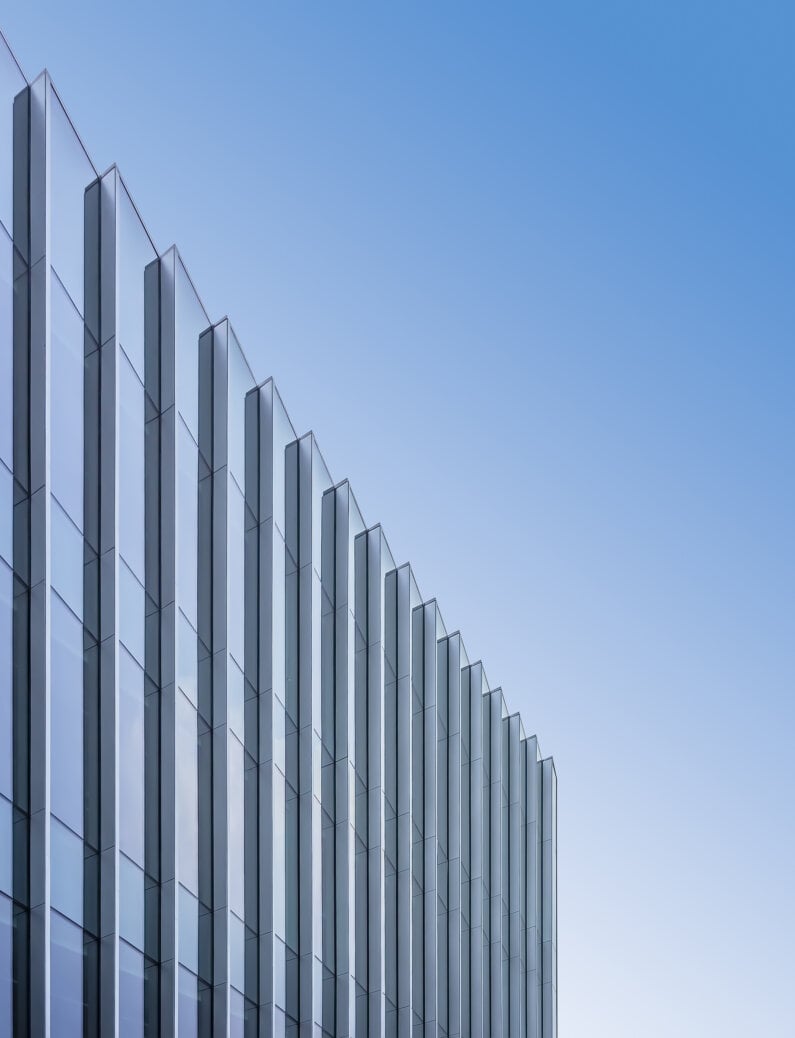 Glass facade of a tall building featuring geometric lines and sharp angles under a clear blue sky, highlighting its sleek architectural design.
