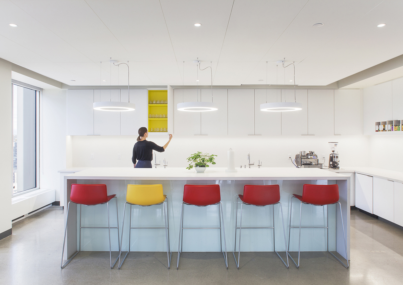 Clean white kitchen with red and yellow bar chairs and woman opening cabinet to show pop of yellow inside