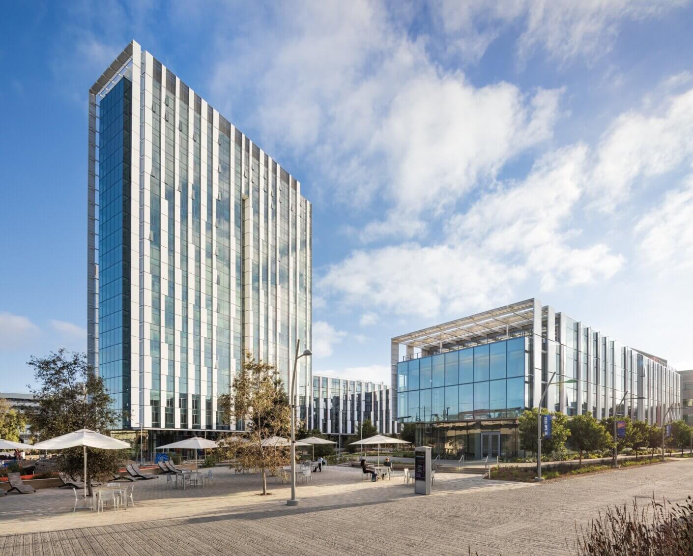 Modern glass office buildings with outdoor seating and trees under a blue sky.