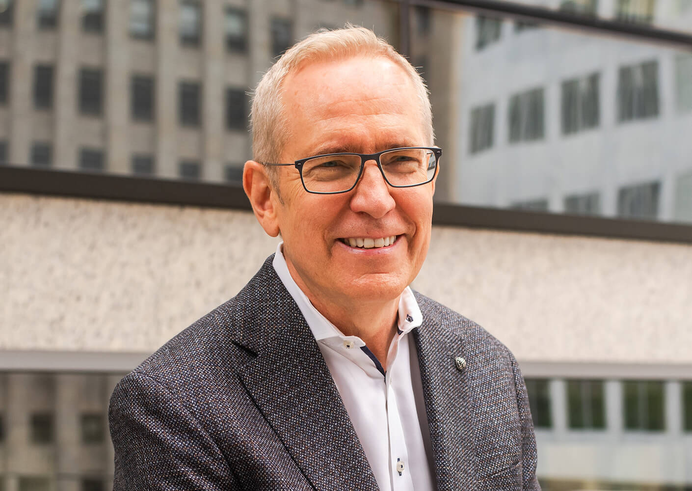 Smiling middle-aged man in glasses wearing a blazer and white shirt stands outside a building with reflective windows.