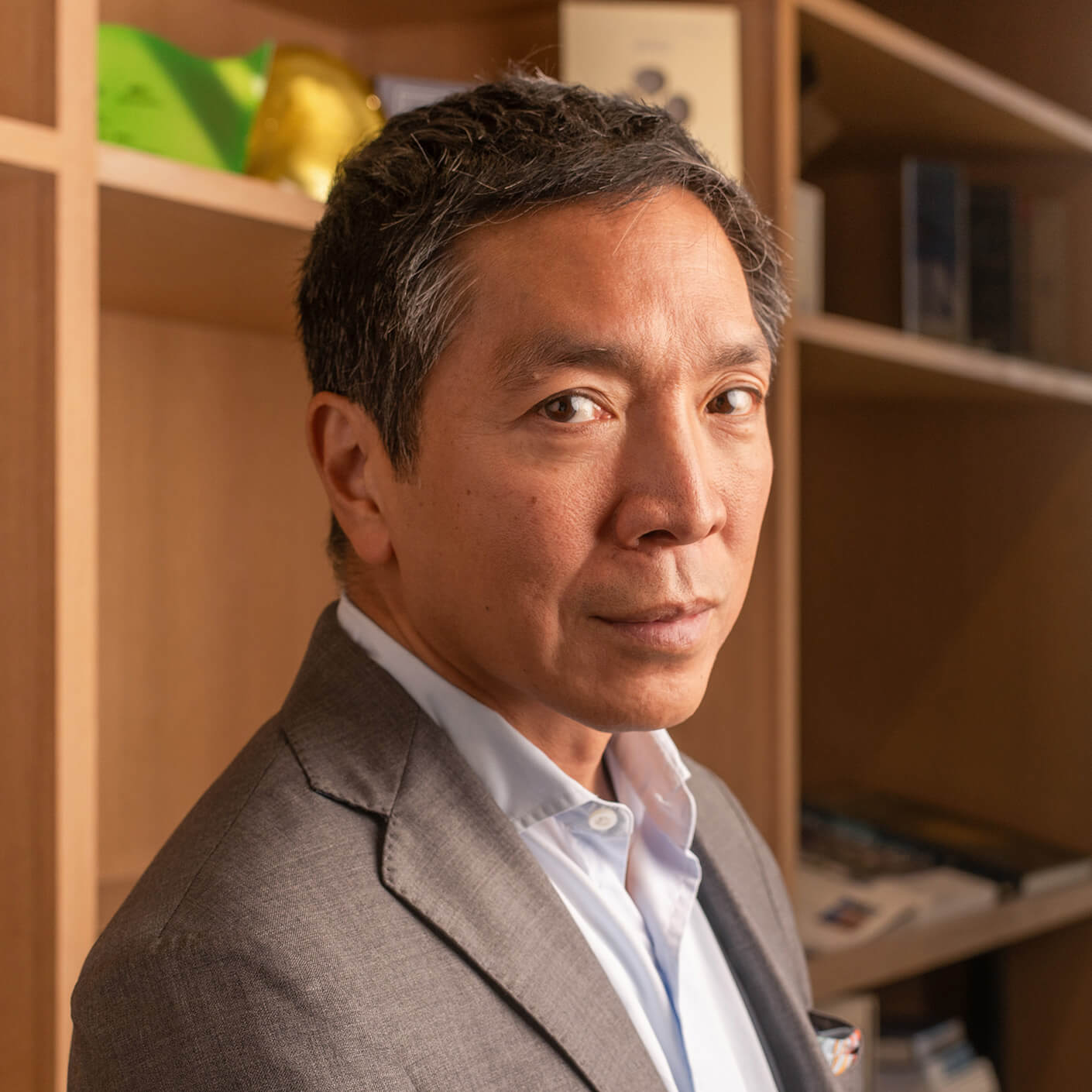 Business professional with short dark hair, wearing a gray suit, poses in an office setting with bookshelves in the background.
