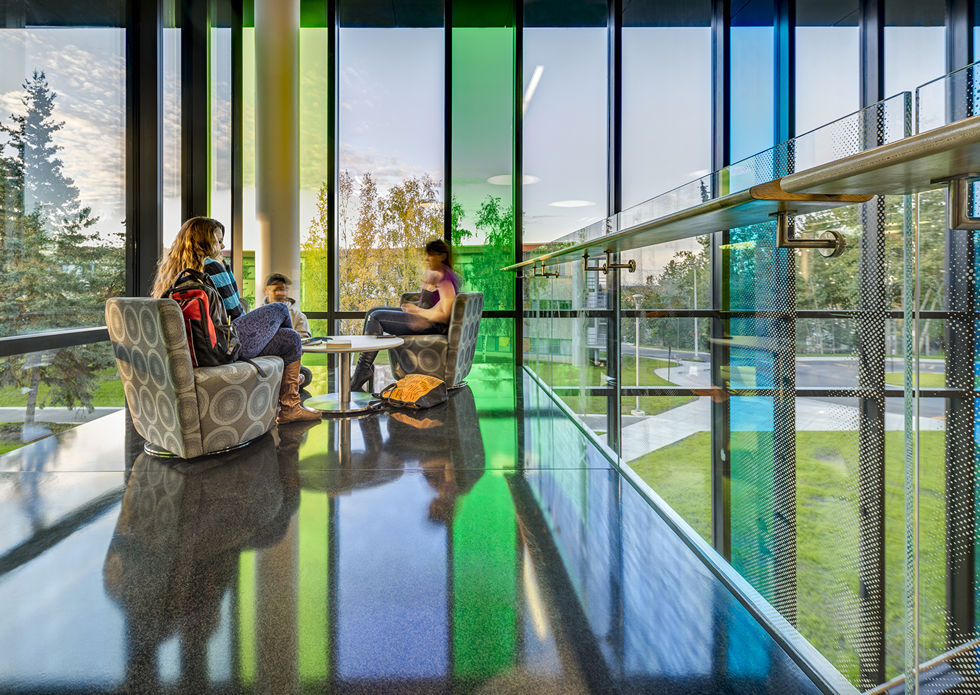 Students sitting and talking in soft seating with bright panes of class behind and views to outside.