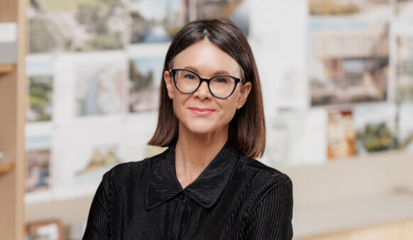 A professional woman with shoulder-length hair and glasses smiles, standing in front of design sketches and architectural plans.