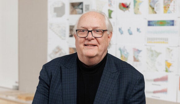 Portrait of a man with glasses and gray hair, wearing a black turtleneck and blue plaid blazer, smiling in front of a wall of maps and diagrams.
