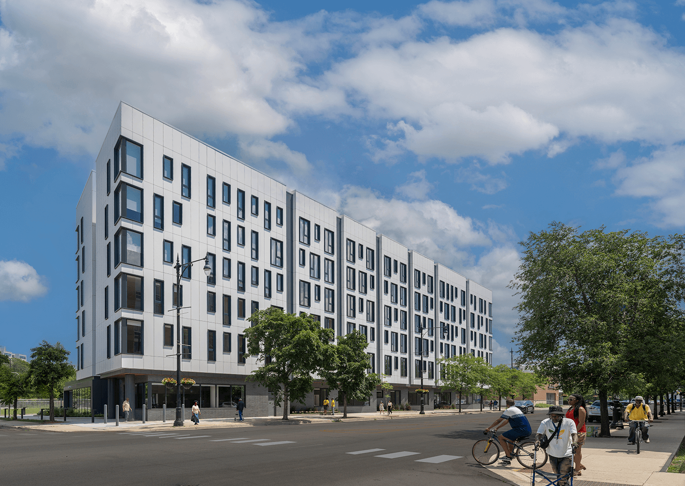 Modern white building with trees and bicyclists on sunny urban street.