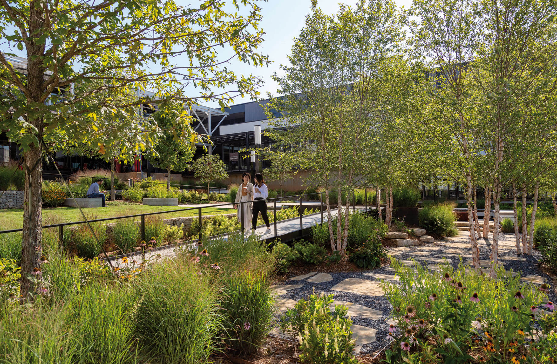 Modern office garden featuring trees, flowers, and two people walking on a bridge.