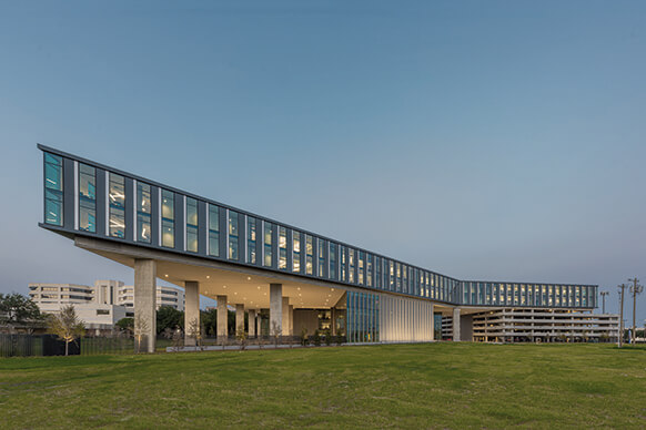 Modern, horizontal building with glass facade, partially elevated on columns over a green lawn at dusk.