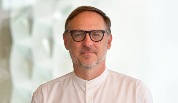 Portrait of a man with glasses and a beard, wearing a light-colored shirt, standing against a blurred background.