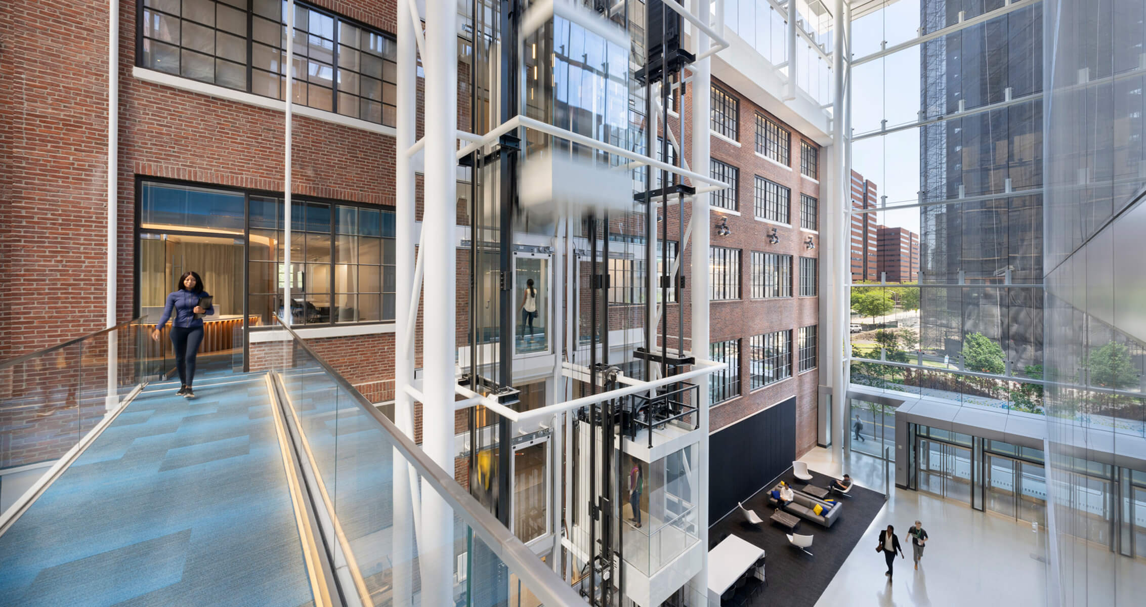 Interior view of a multi-level building showcasing glass elevators, brick walls, and a walkway with people navigating the space.