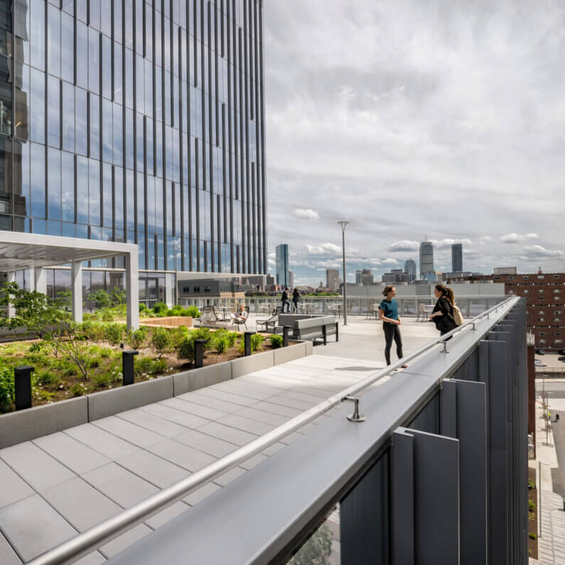 Two people converse on a rooftop terrace with greenery, seating areas, and city skyline views in the background. Cloudy sky overhead.