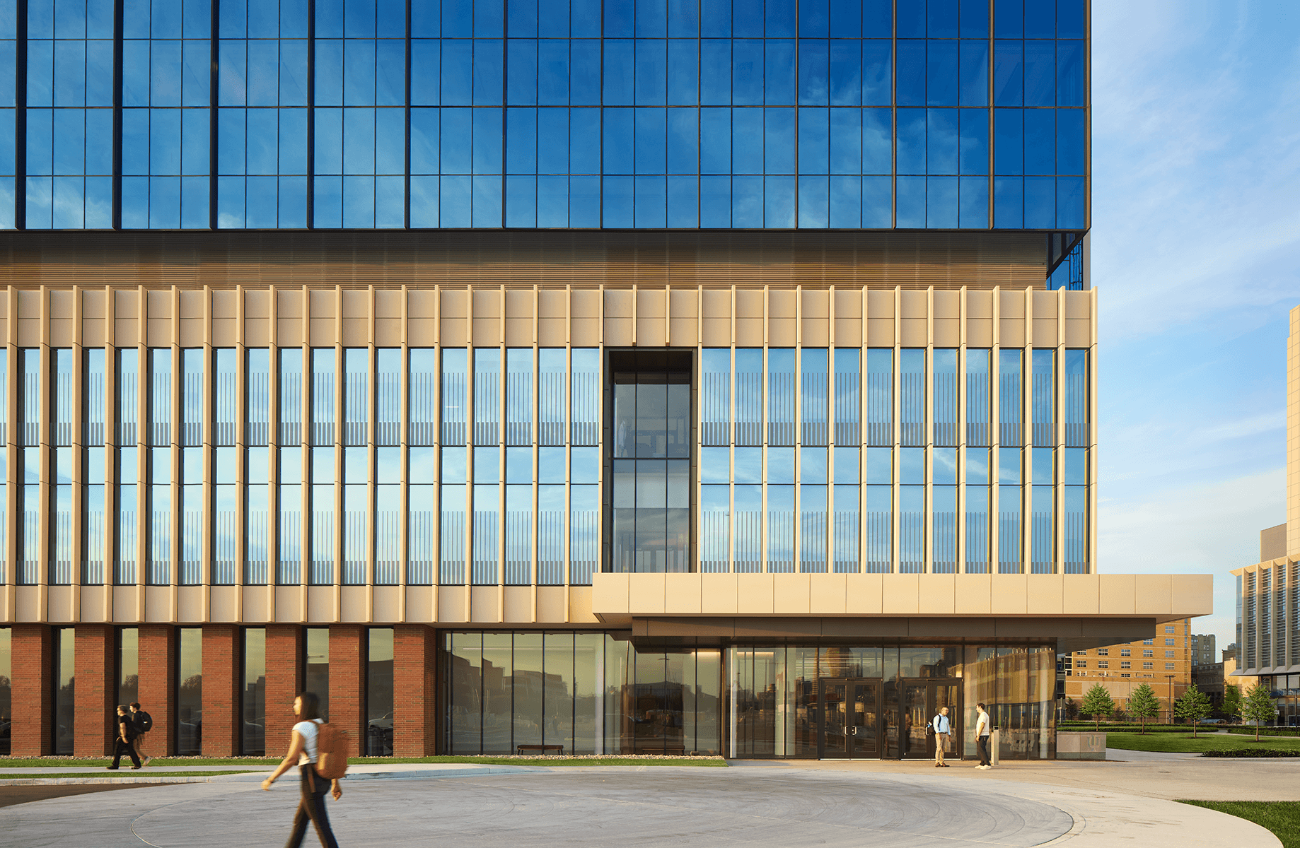 Architectural facade of a building featuring large glass windows, horizontal bands, and brick accents, with pedestrians walking in front.