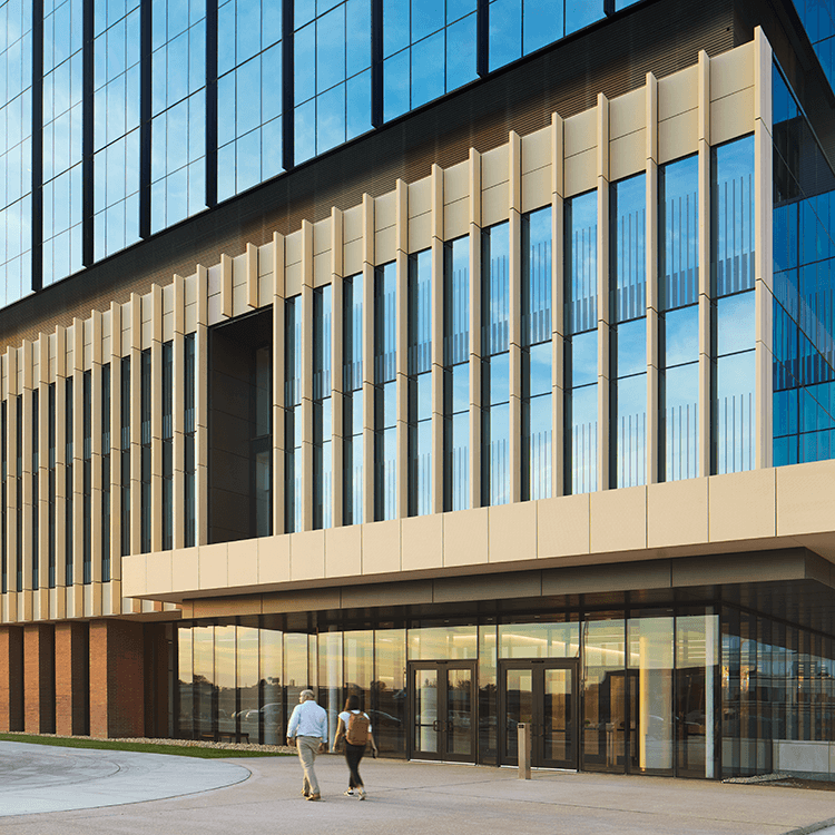 Two people walk towards a building with large glass windows and vertical facade elements, surrounded by open space and evening light.