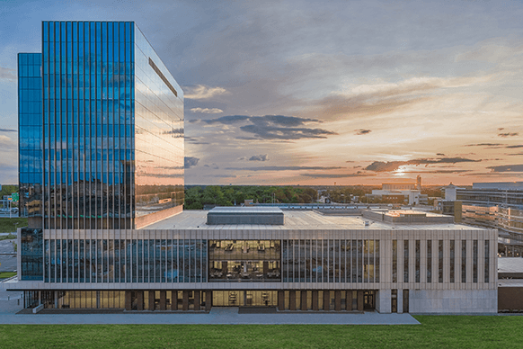 Aerial view of a sleek, multi-story building with glass façade reflecting a sunset, surrounded by green space and other structures.