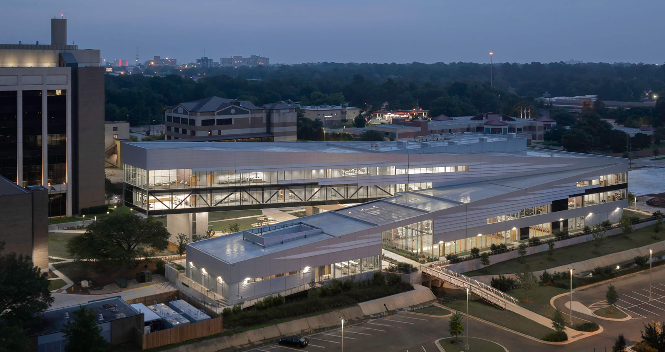 Modern building with glass facade at night, surrounded by greenery and cityscape.