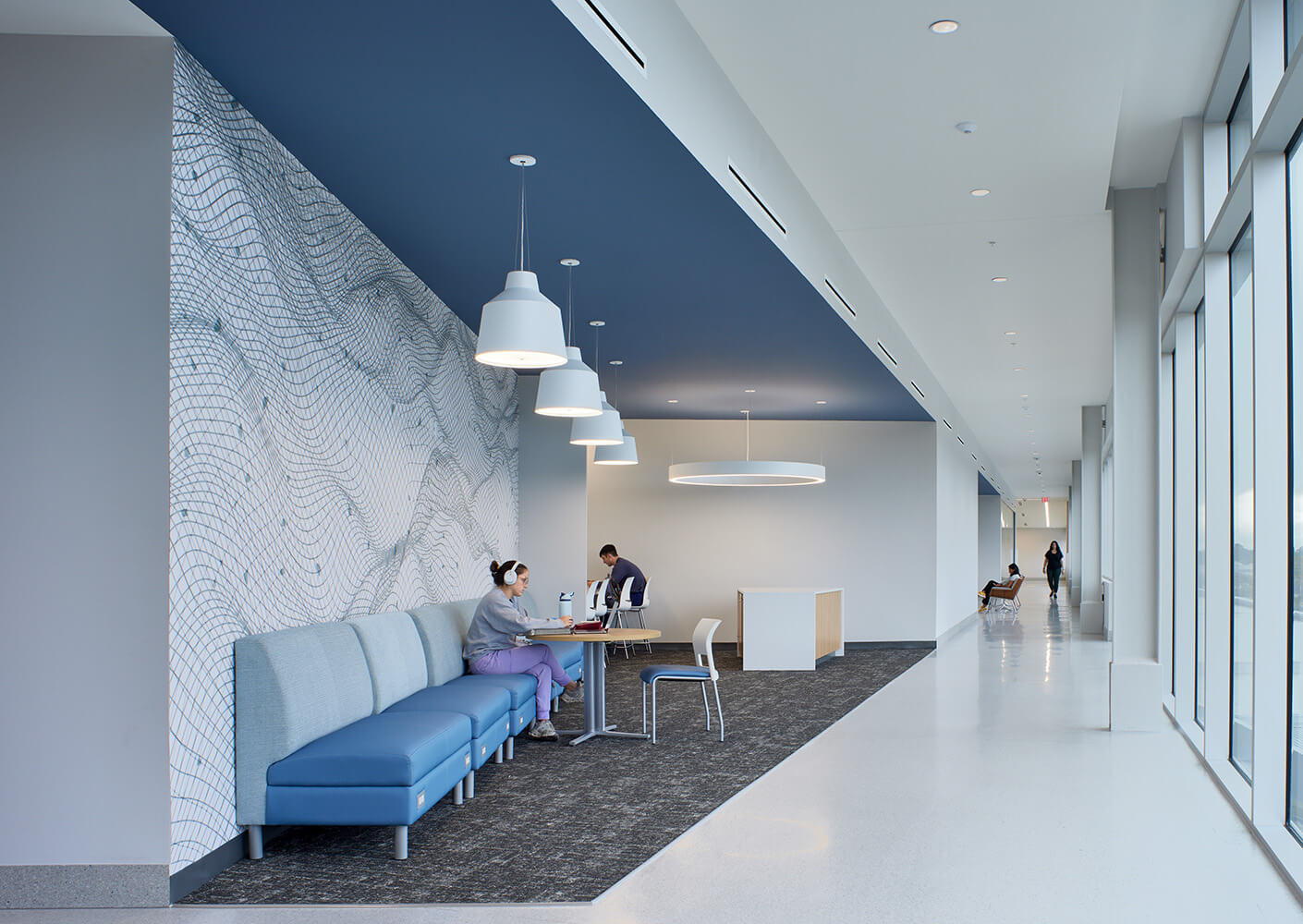 Bright workspace niche along a hallway with people working at tables near large windows.
