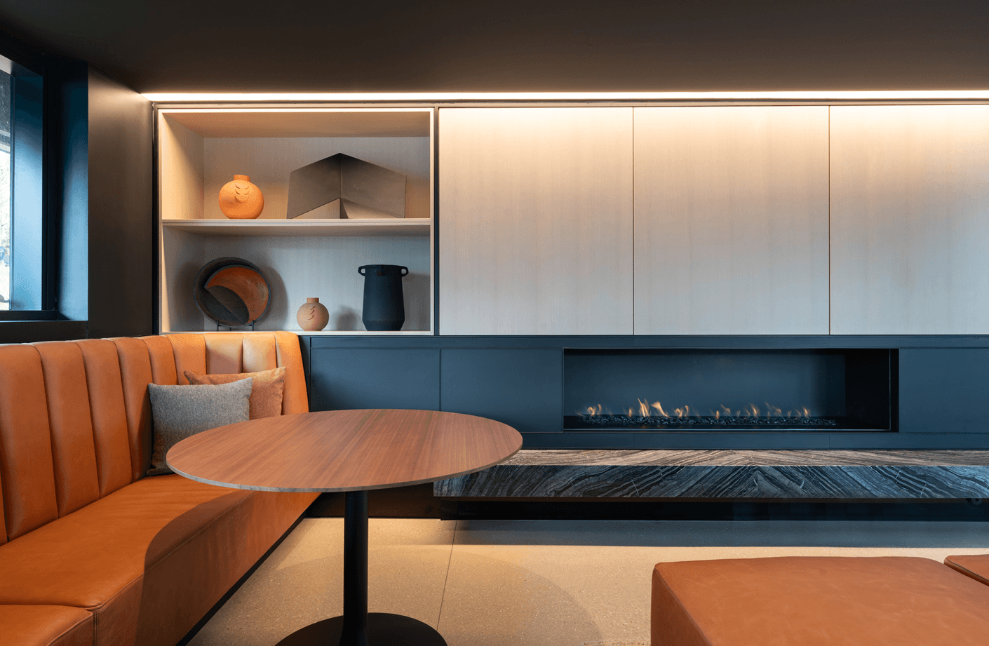 Cozy lounge area featuring an orange leather bench, circular wooden table, and decorative shelves with pottery and a fireplace.