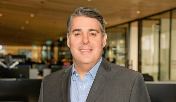 Professional man in a suit smiles in an office space with glass walls and workstations in the background, conveying a business environment.