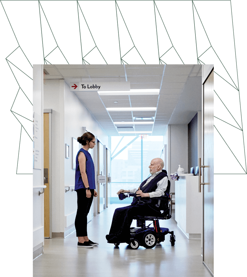 a doctor with muscular distrophy sits in his wheelchair and talks with a woman in the hallway of a medical facility