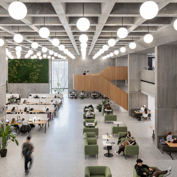 A second story view of the atrium, showing a concrete room illuminated by white orb lights. A wooden staircase and information desk as well as a moss wall stand out against the hues of the wall.