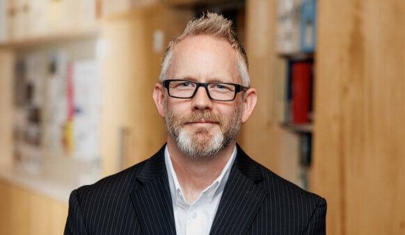 Business professional with glasses and a beard, wearing a pinstripe suit, poses for a portrait in an office setting with blurred background.
