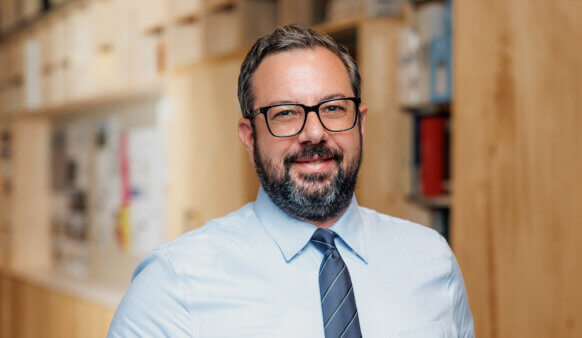 Portrait of a smiling man with glasses and a beard, wearing a light blue shirt and tie, set against a backdrop of wooden surfaces and design materials.