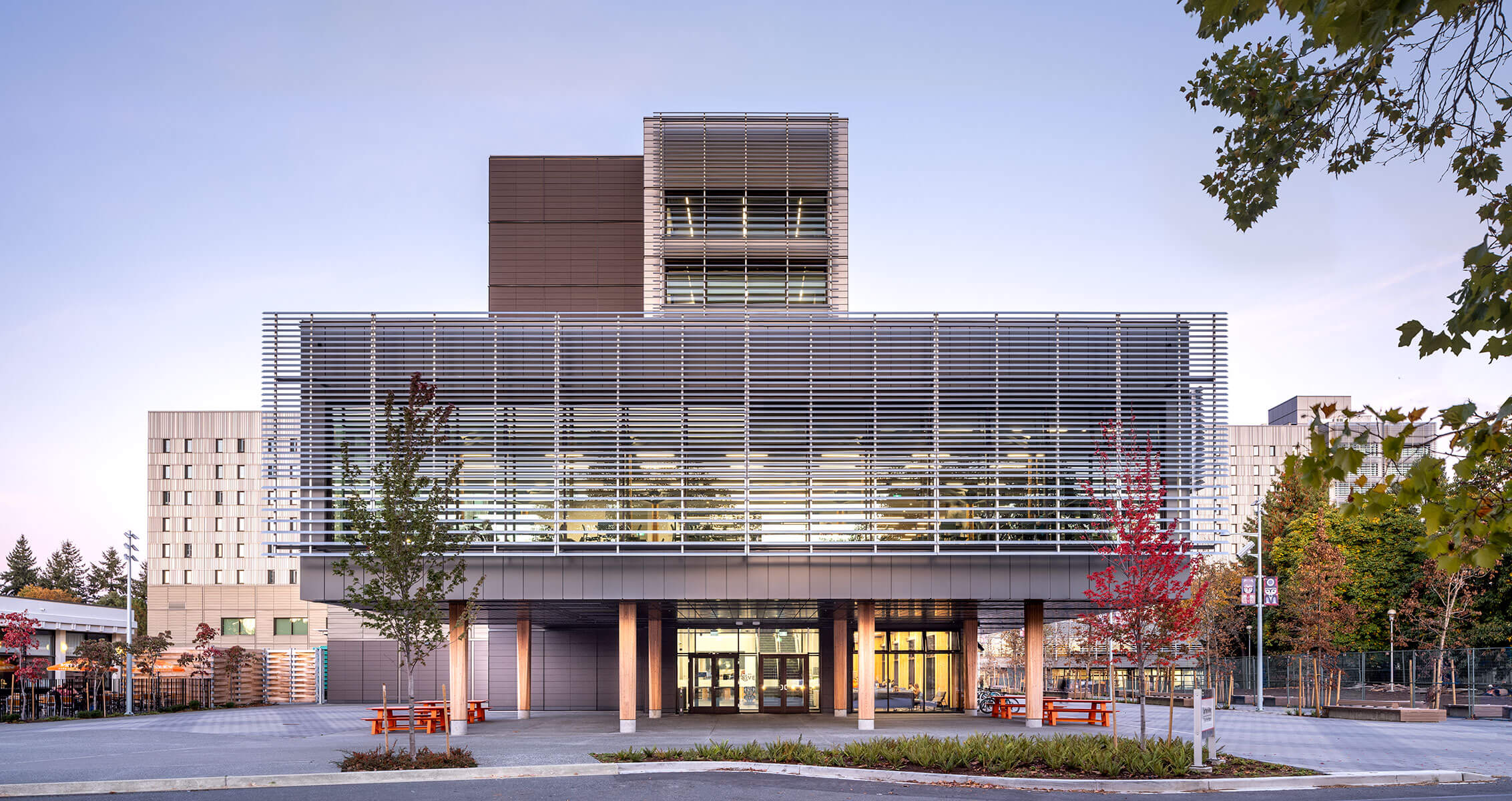 Dusk photograph of the entrance of Cheko’nien House with multipurpose room above and residential tower in the background.