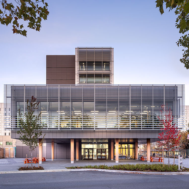 Dusk photograph of the entrance of Cheko’nien House with multipurpose room above and residential tower in the background.