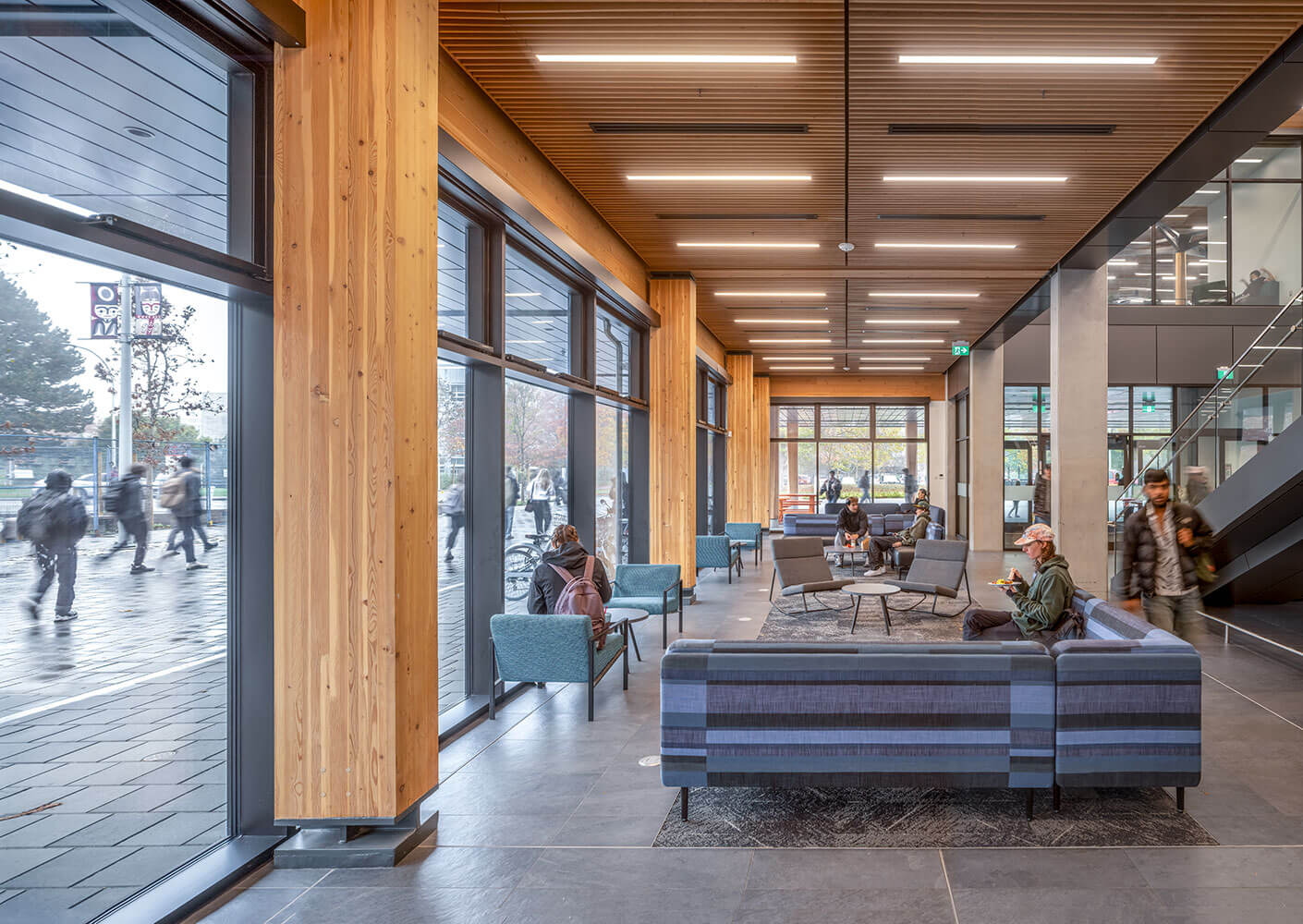 Photograph of some lounge seating in the servery of Cheko’nien House with student study and eating. The fully glazed common areas provide views to the Campus Greenway with students walking outside.