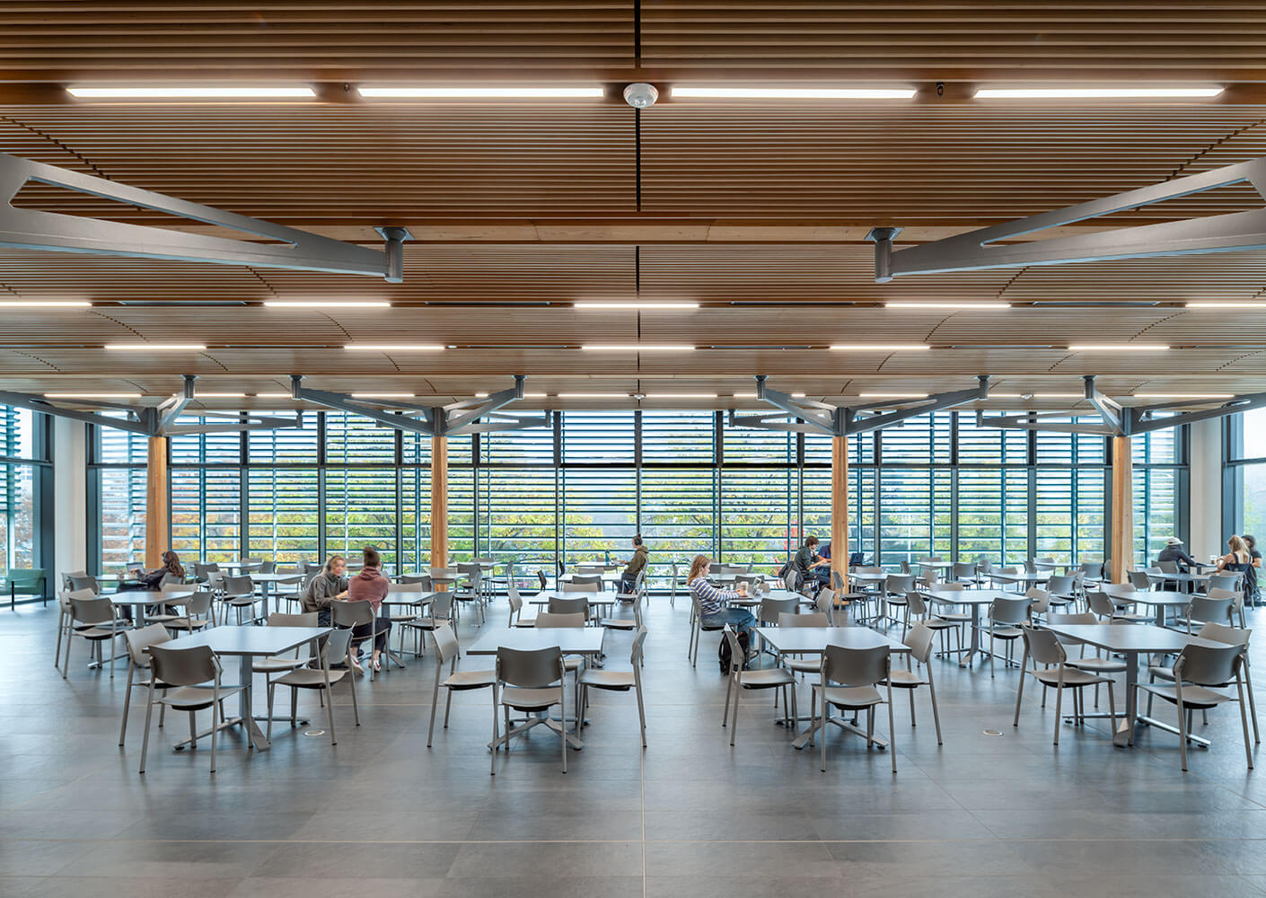 Photograph within the multipurpose room looking west towards Ring Road. The room is configured with tables chairs with several students working. The wood ceiling with integrated lighting and large wooden columns are featured.