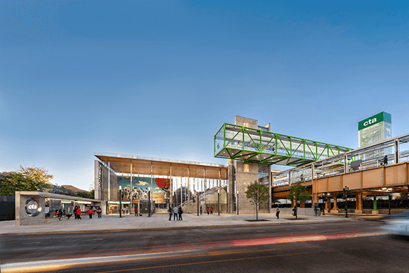 Modern urban transit hub with green walkway, glass architecture, and motion-blurred traffic against a clear sky.