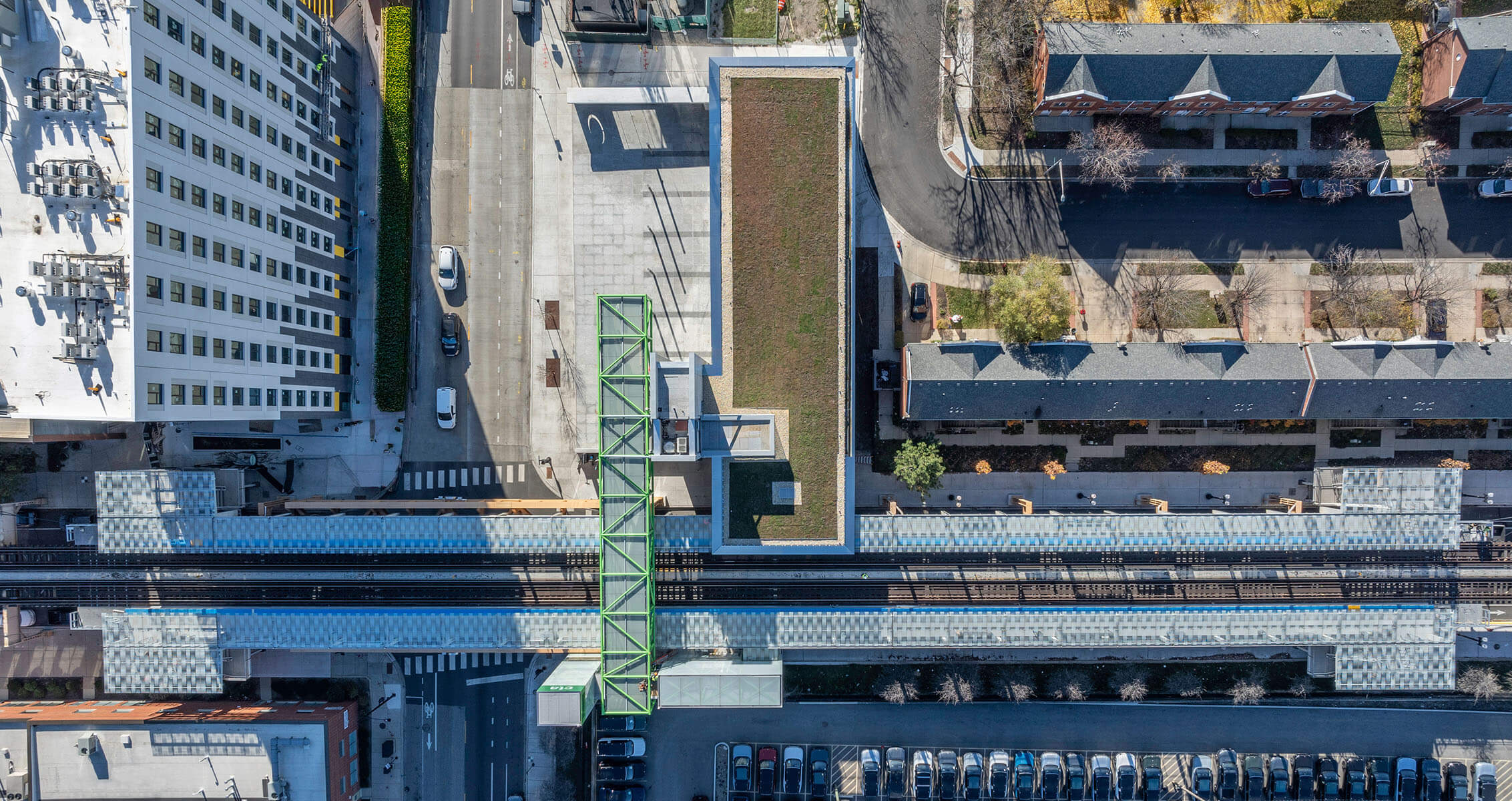 Aerial view of urban street and railway station with green roof and parked cars on a sunny day.