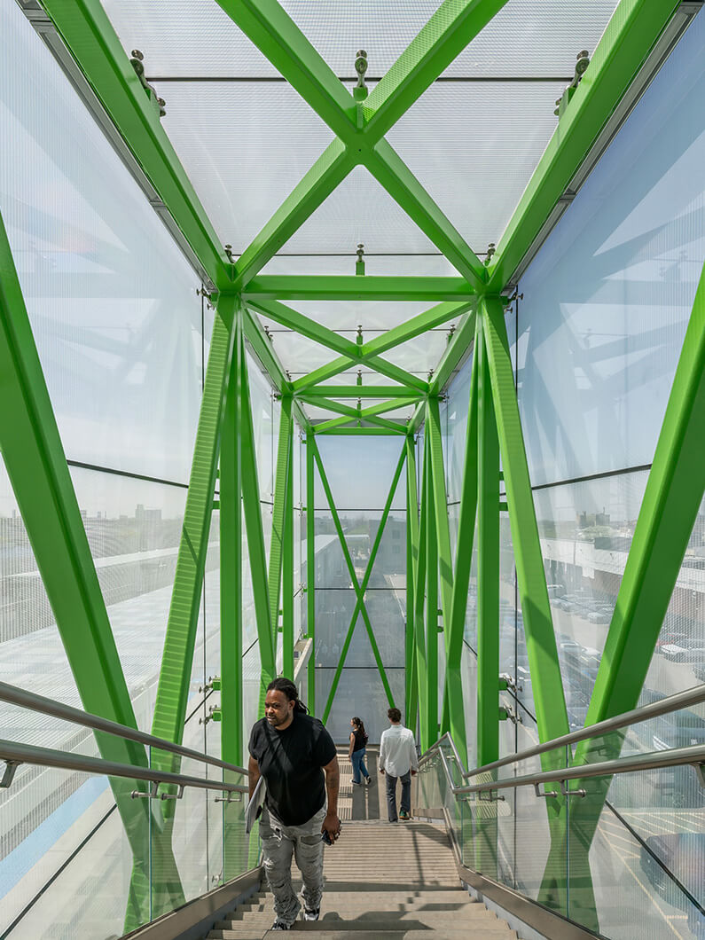 Man ascending stairs in a green metal-framed pedestrian bridge with clear panels and urban views.