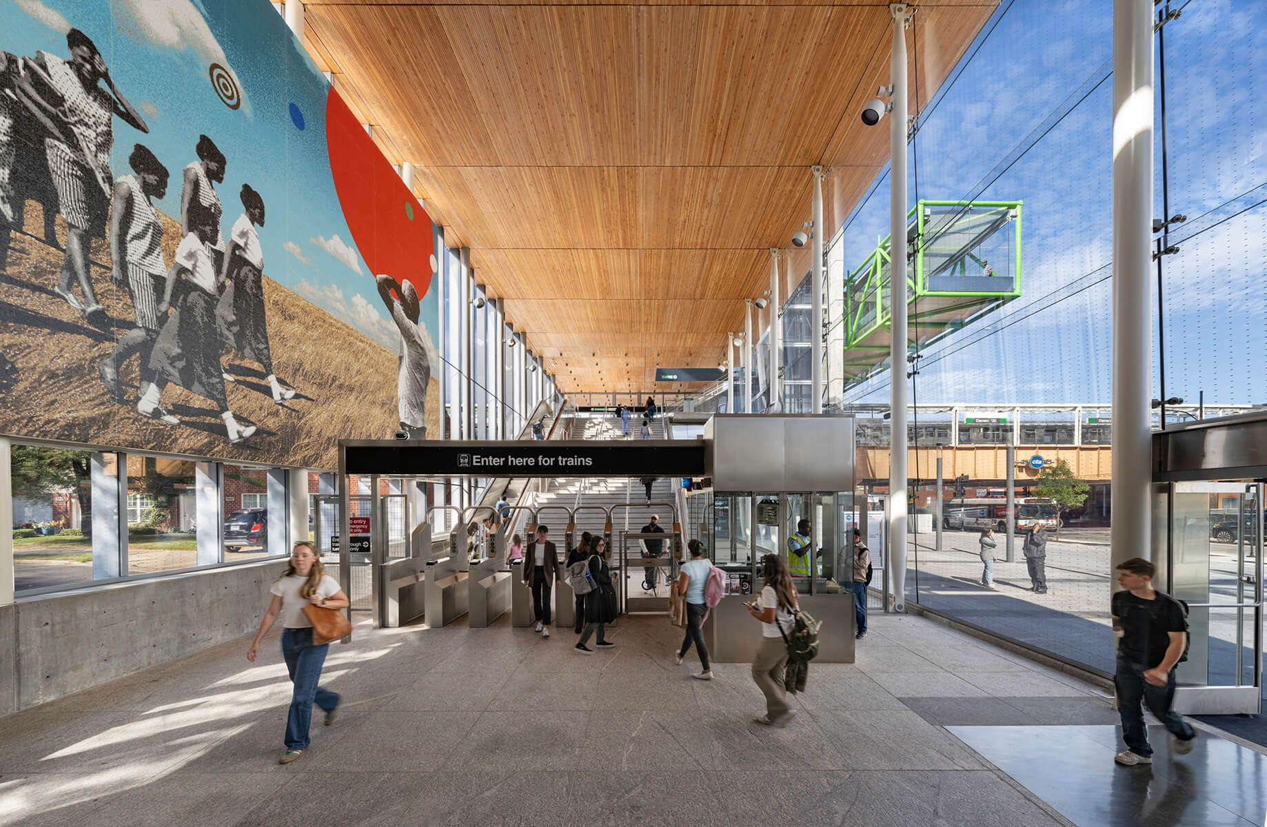 Modern train station entrance with people passing through. Large mural and glass walls create an open, airy atmosphere.