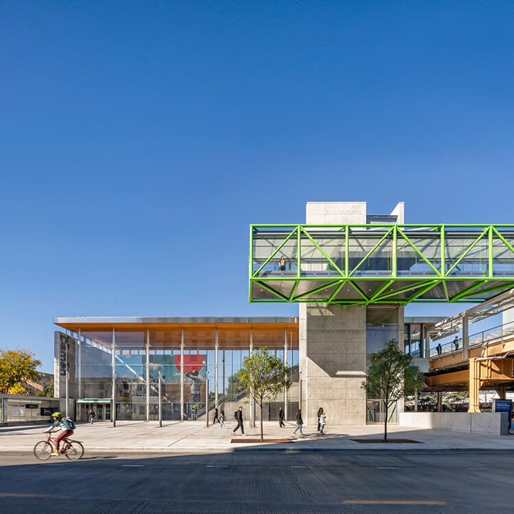 Modern train station with green skywalk, cyclists, and pedestrians under a clear blue sky.