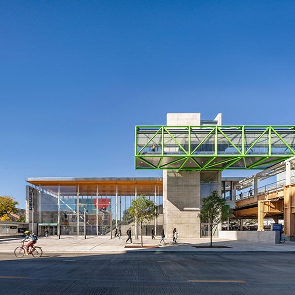 Modern transit station with glass facade, green overhead walkway, and commuters under a clear blue sky.
