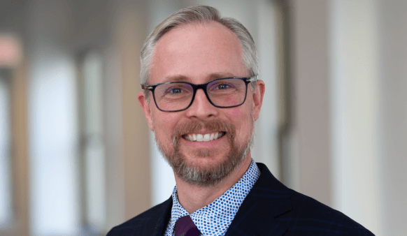 Professional headshot of a smiling man with glasses in a suit and tie.