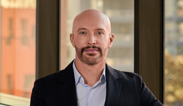 Portrait of a man with a shaved head and a beard, wearing a blazer and shirt, standing by a window with city views in the background.
