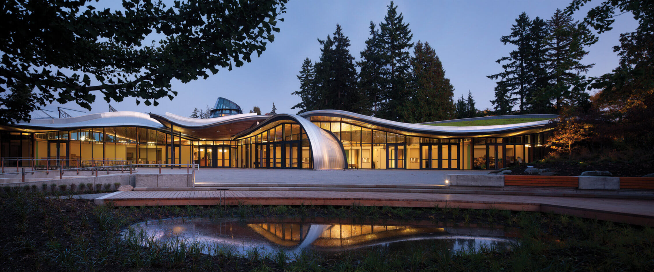 An organic, curving wood and metal building at dusk, across a pond