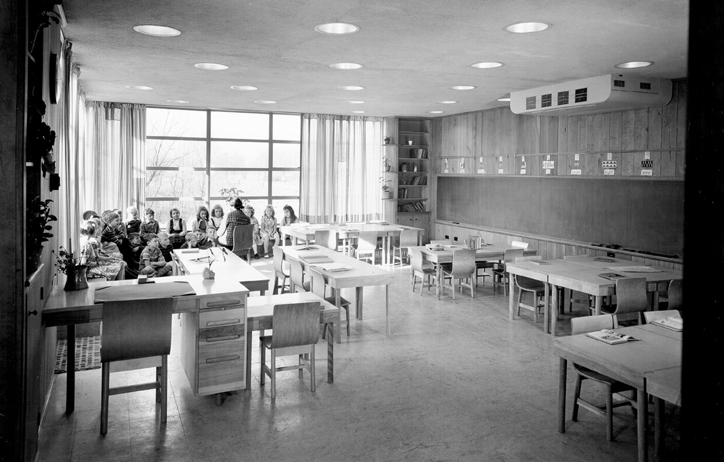 Historical photo of children sitting with their teacher in a daylit classrom