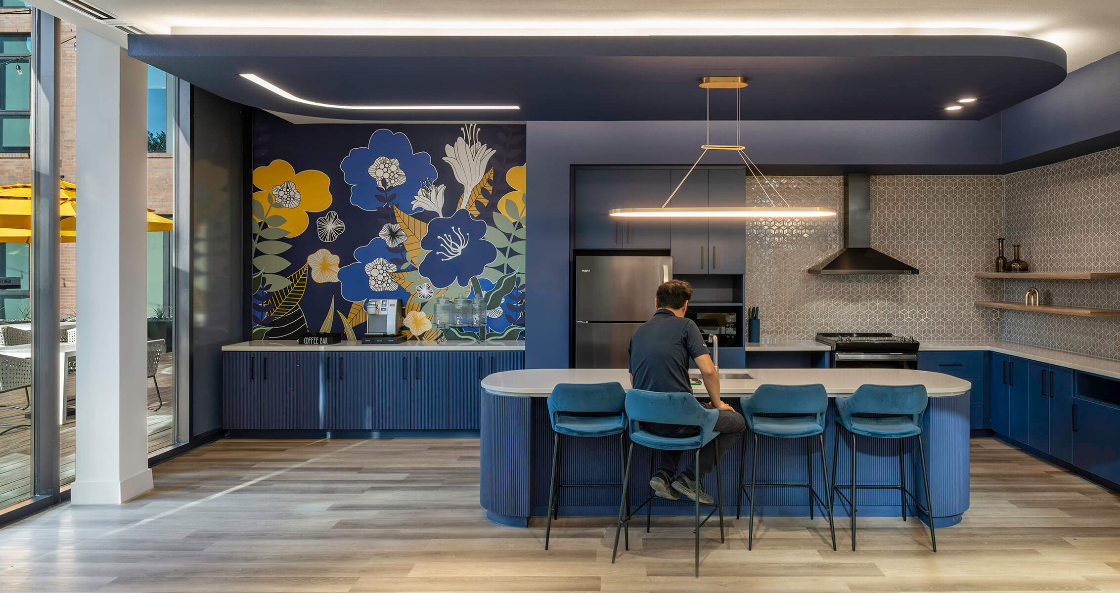 A man sits next to a floor-to-ceiling window in a navy blue community space featuring a floral mural