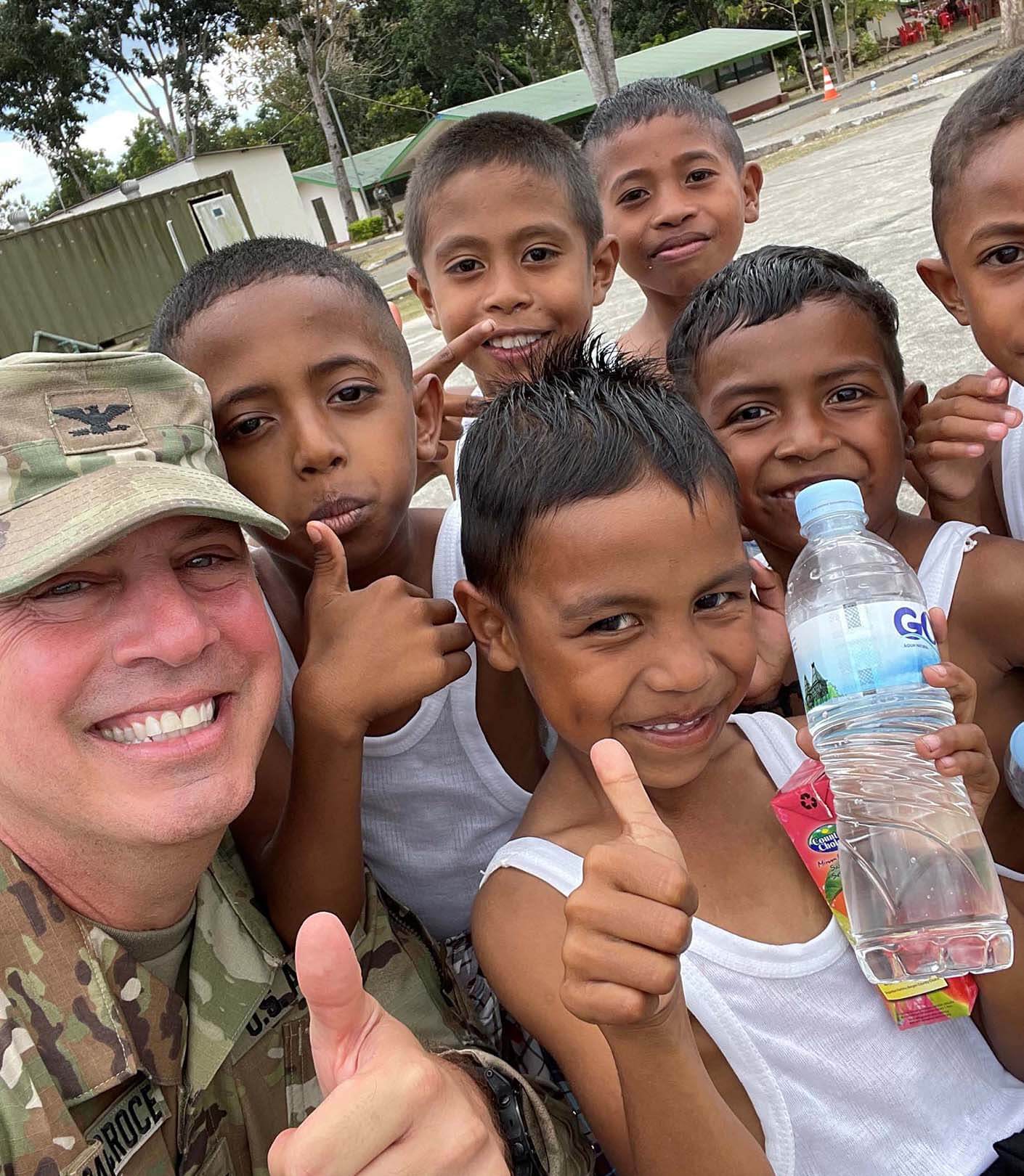 Rudy in army uniform smiling with a group of children in Timor-Leste, giving a thumbs up